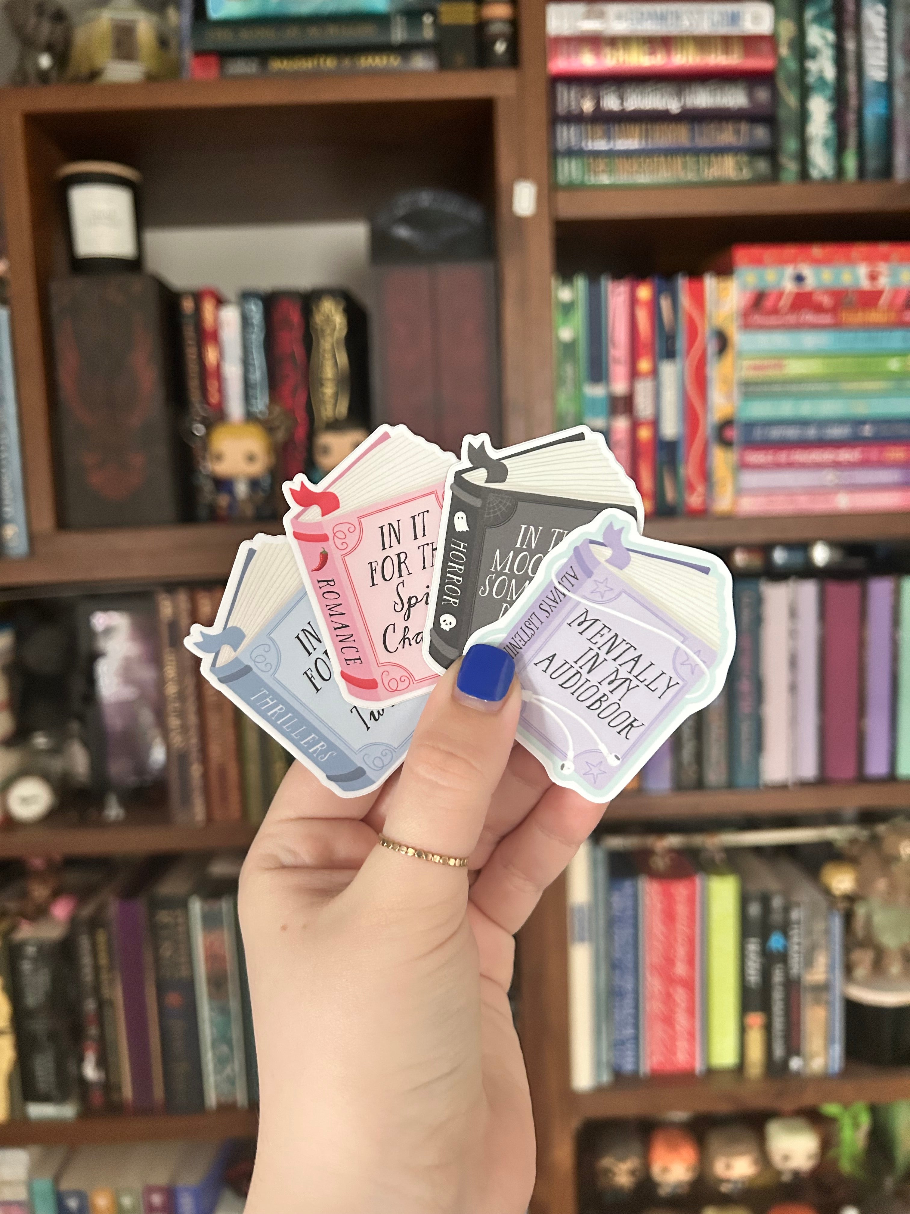 A hand with blue nail polish holds a fanned-out selection of die-cut stickers featuring different color book stickers. 
