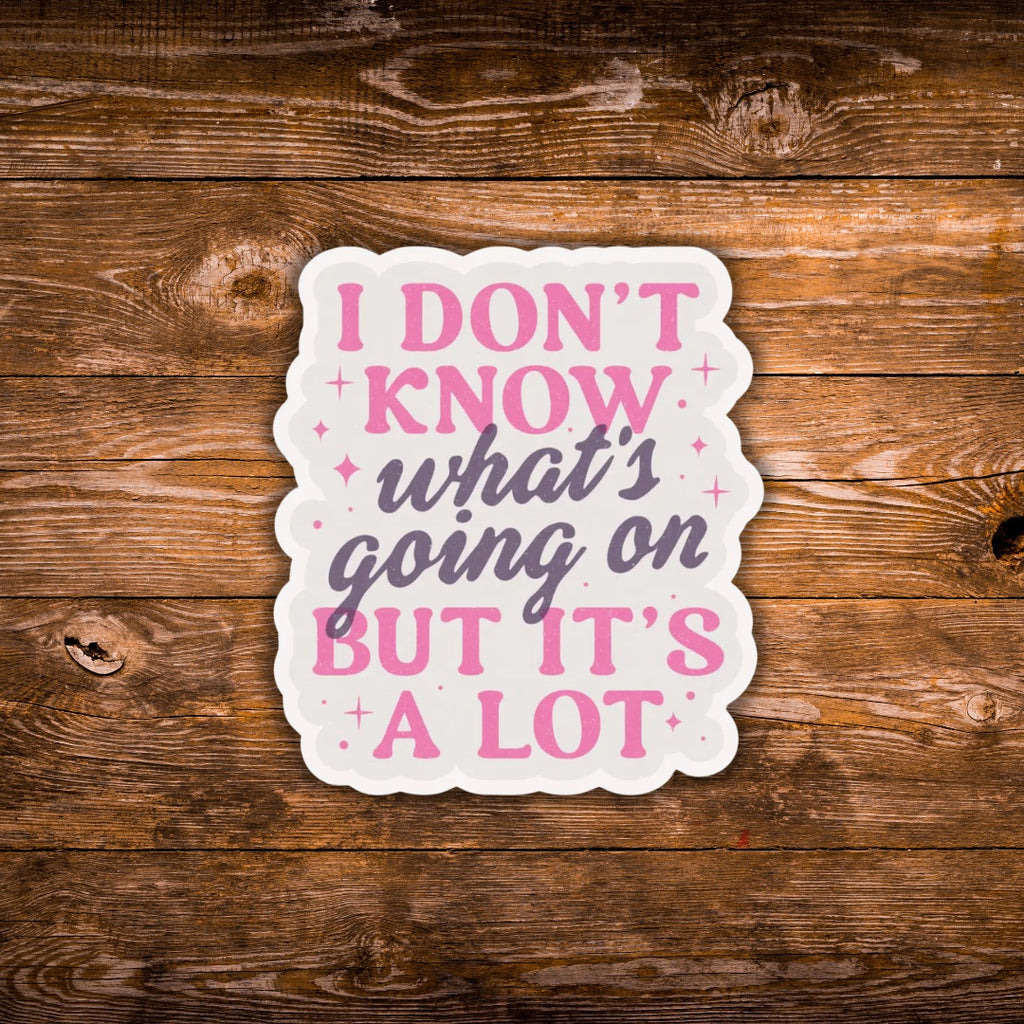 A scalloped die-cut sticker with the text 'I DON'T KNOW what's going on BUT IT'S A LOT' in pink and dark gray lettering on a white background, displayed on a rustic brown wooden surface.