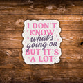 A scalloped die-cut sticker with the text 'I DON'T KNOW what's going on BUT IT'S A LOT' in pink and dark gray lettering on a white background, displayed on a rustic brown wooden surface.