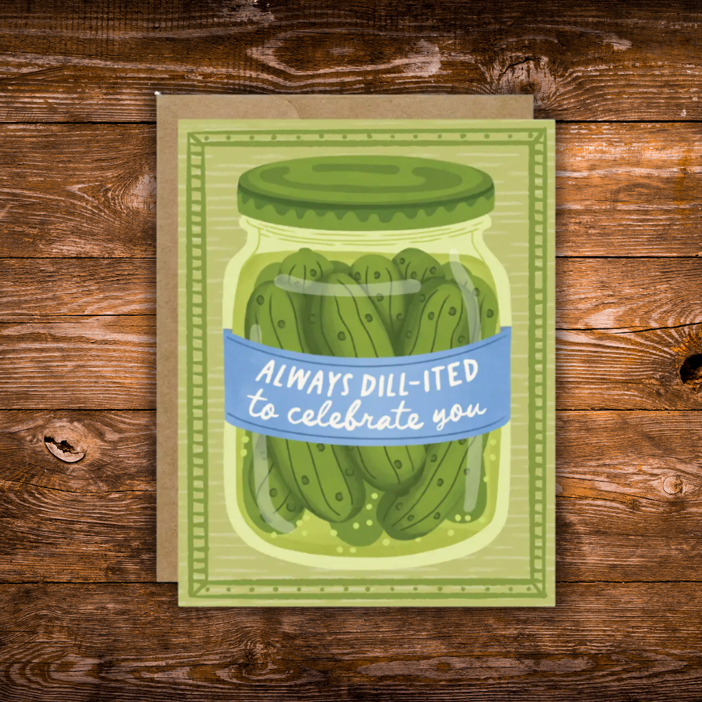 A square greeting card featuring an illustrated jar of dill pickles with a blue banner reading 'ALWAYS DILL-ITED to celebrate you,' placed on a brown kraft envelope over a rustic wooden surface.