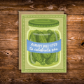 A square greeting card featuring an illustrated jar of dill pickles with a blue banner reading 'ALWAYS DILL-ITED to celebrate you,' placed on a brown kraft envelope over a rustic wooden surface.