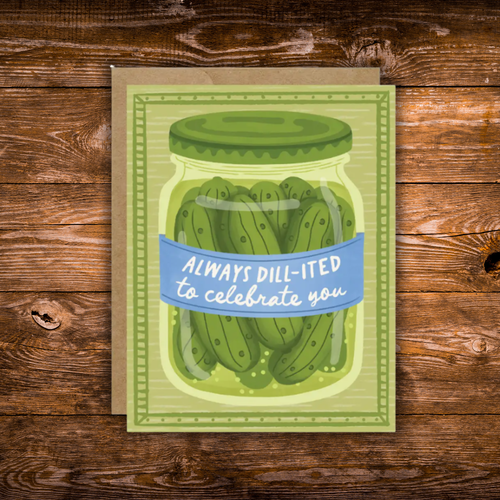 A square greeting card featuring an illustrated jar of dill pickles with a blue banner reading 'ALWAYS DILL-ITED to celebrate you,' placed on a brown kraft envelope over a rustic wooden surface.