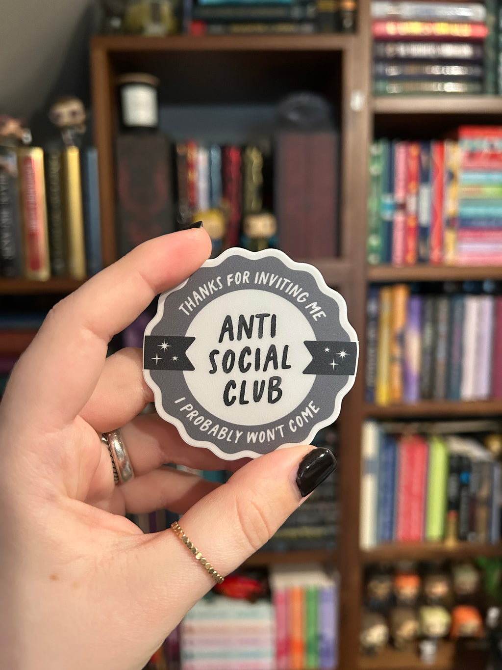 A hand with black nail polish holds a scalloped die-cut sticker that reads 'THANKS FOR INVITING ME I PROBABLY WON'T COME- ANTI SOCIAL CLUB' against a blurred background of bookshelves.