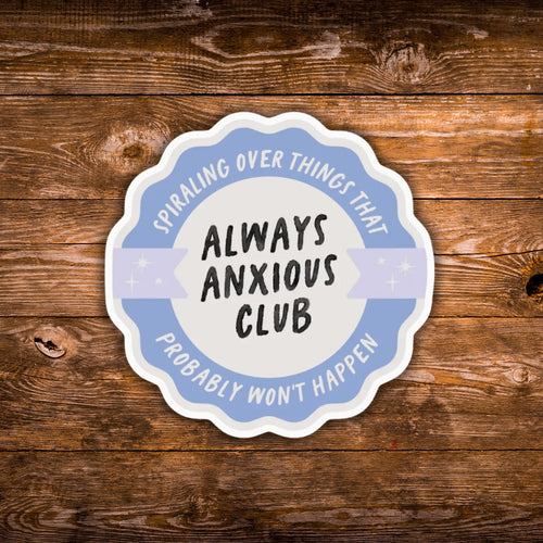 A scalloped die-cut sticker with the text 'ALWAYS ANXIOUS CLUB' in black on a white center, surrounded by a light blue ring reading 'SPIRALING OVER THINGS THAT PROBABLY WON'T HAPPEN,' shown on a rustic brown wood background.