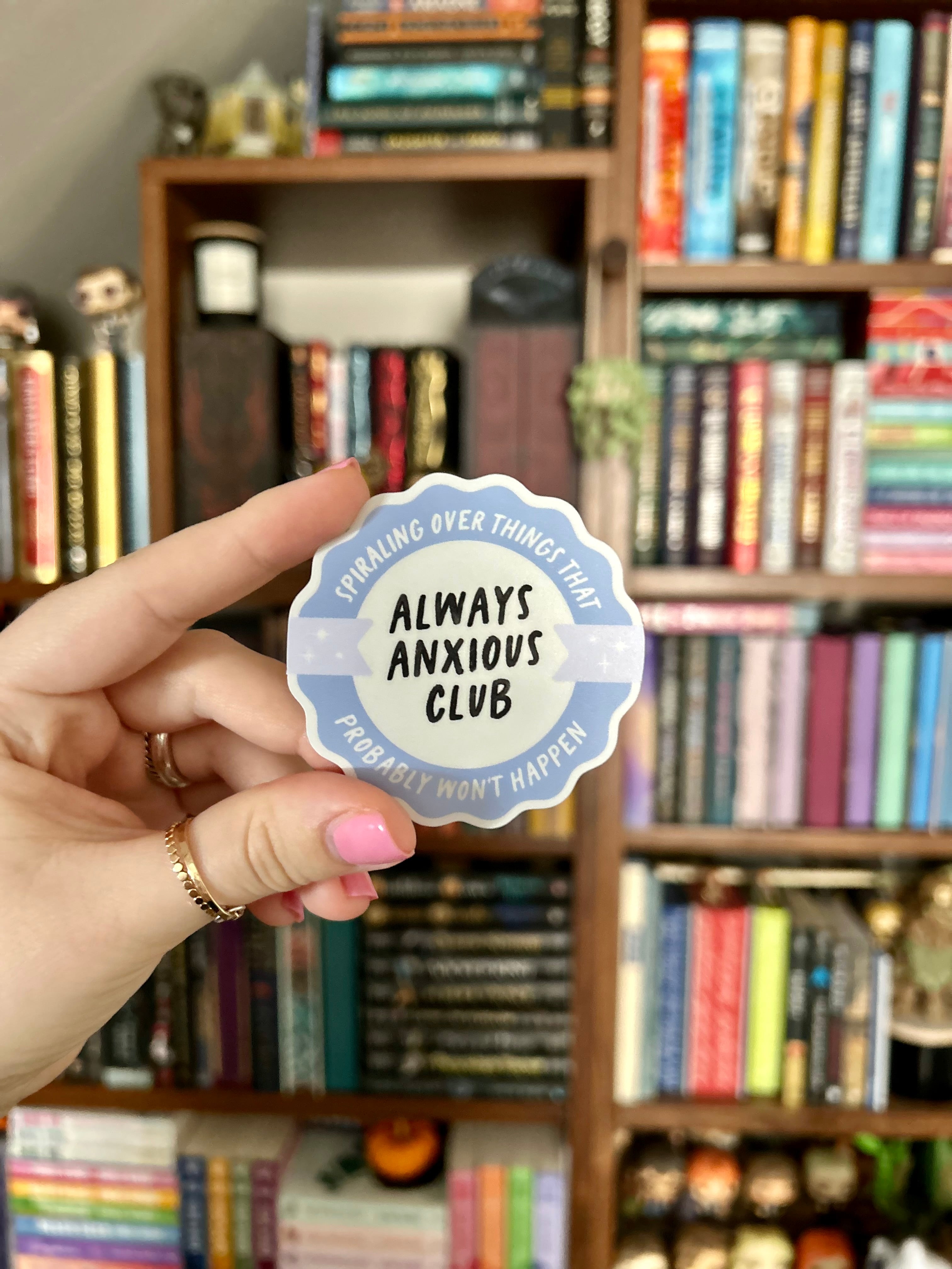 A hand holds a round sticker in front of a blurred bookshelf. The sticker has a light blue border and a white center with the text 'ALWAYS ANXIOUS CLUB' and 'SPIRALING OVER THINGS THAT PROBABLY WON'T HAPPEN'.