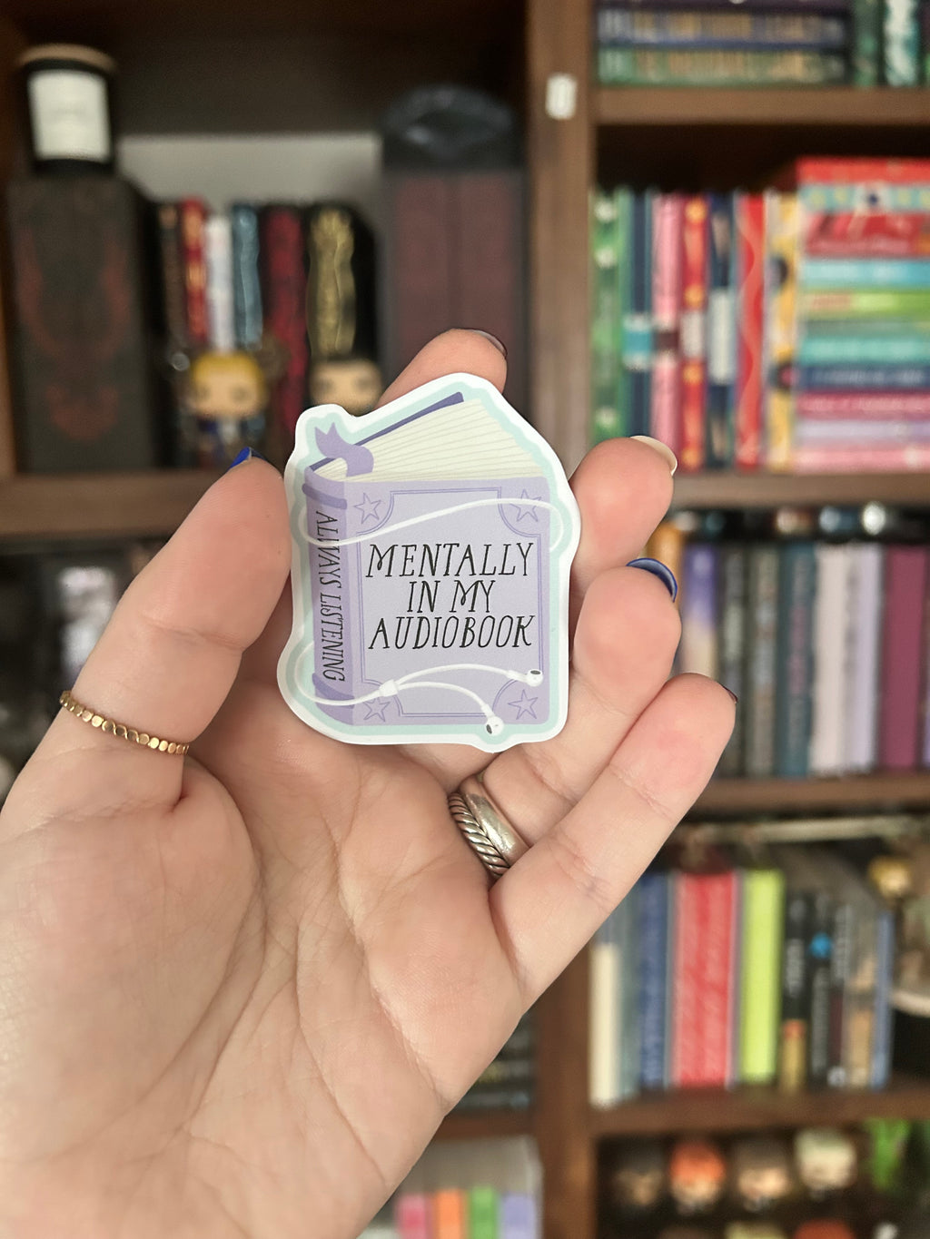 A hand holding a small white and lavender bookmark or card that reads 'MENTALLY IN MY AUDIOBOOK' and 'ALWAYS LISTENING', with a blurred bookshelf in the background.