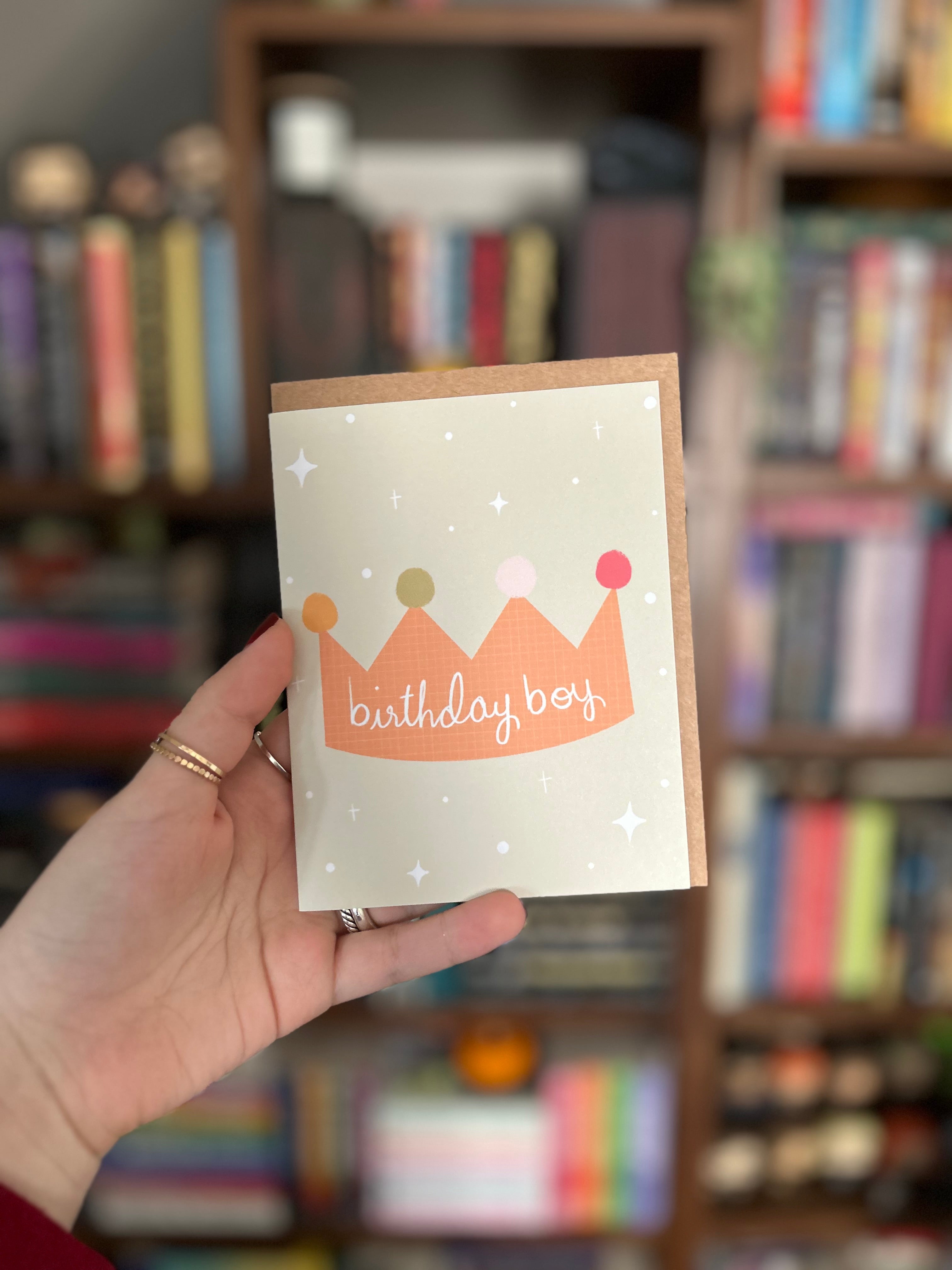 Cute birthday greeting card featuring a colorful illustrated crown with the words "Birthday Boy" on a soft sage green background with scattered stars.