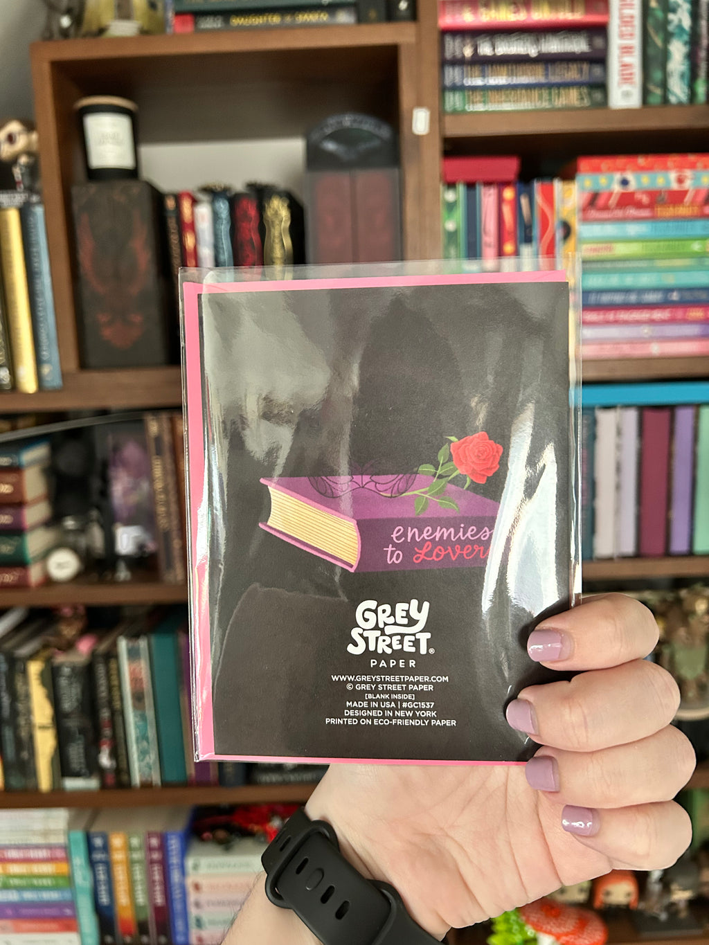 A hand holding up the back of a packaged Grey Street Paper greeting card featuring a black background with a pink book illustration and a red rose, set against a blurred background of bookshelves.