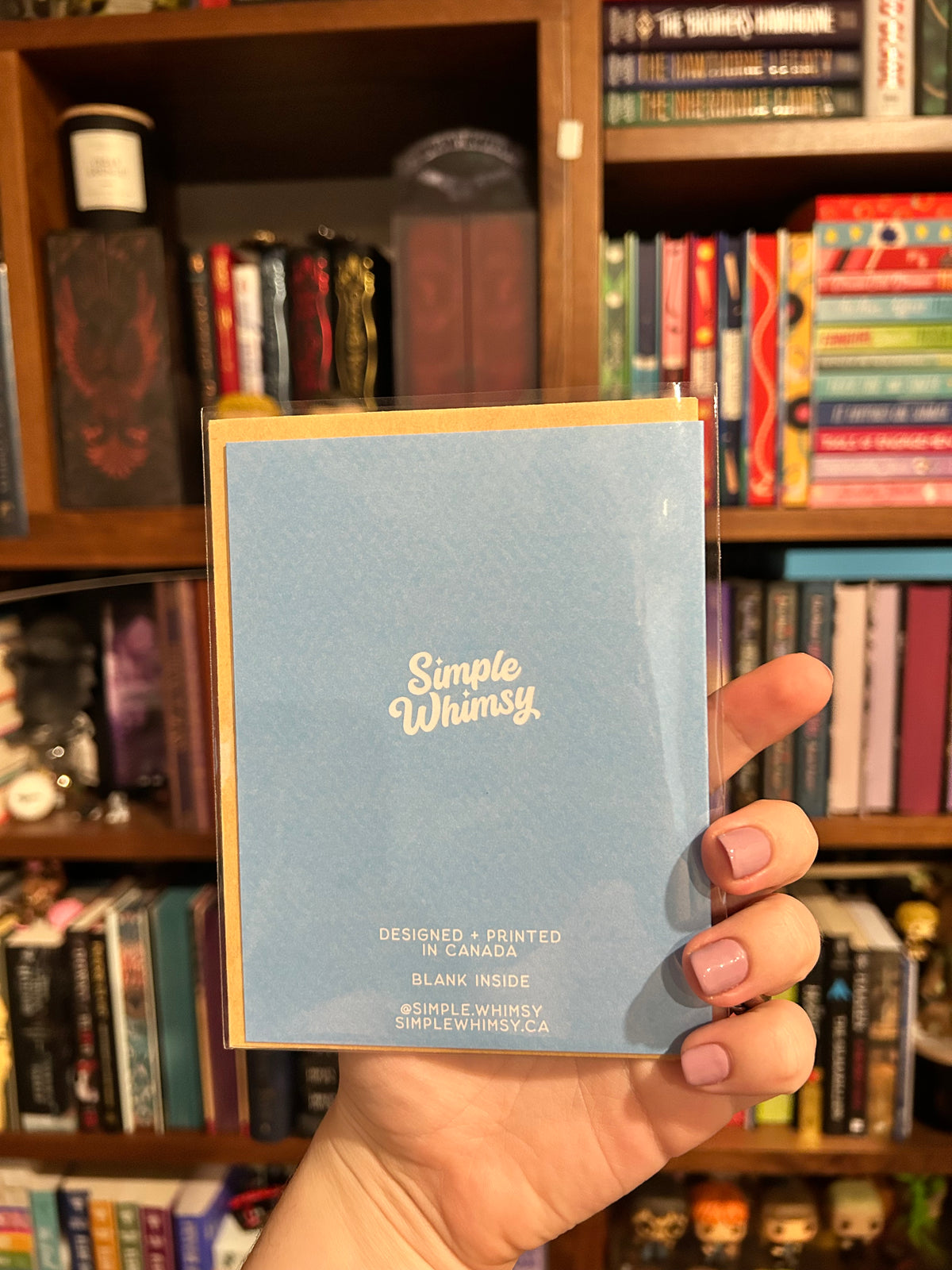 A hand holding a light blue greeting card with white text reading 'Simple Whimsy' in front of a bookshelf filled with books.