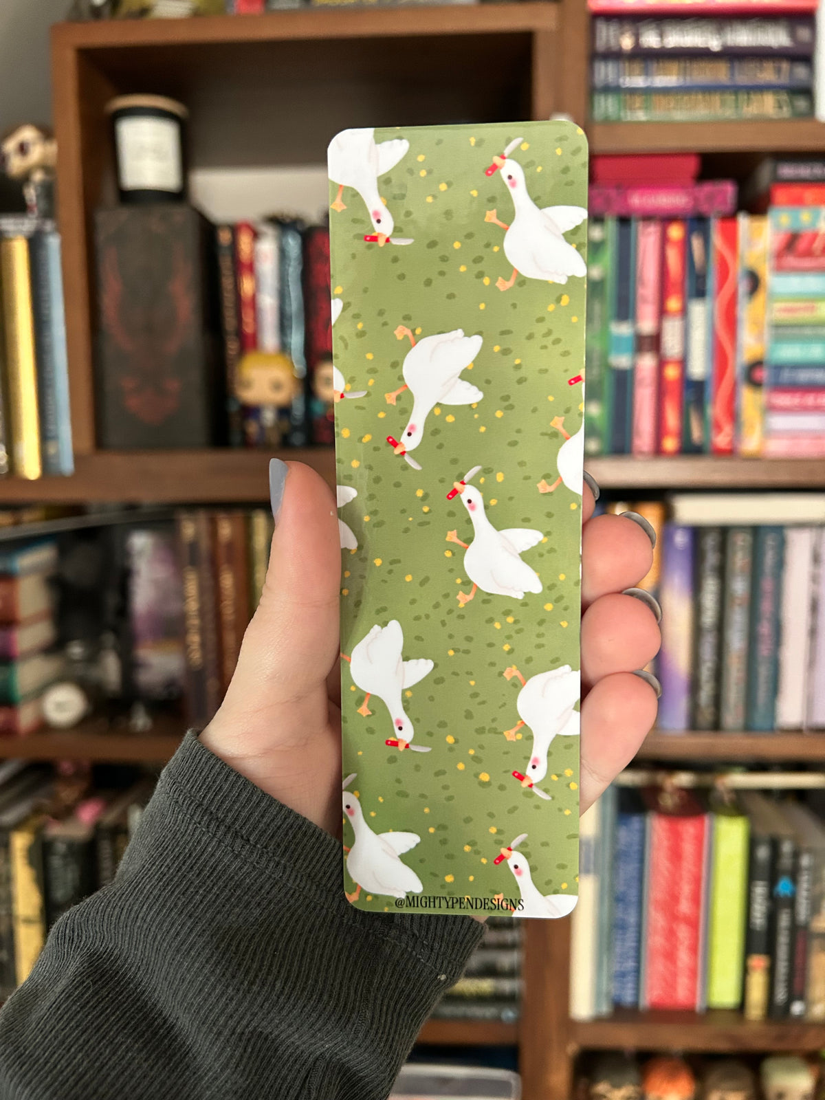 Double-sided goose bookmark featuring a white goose holding a tiny knife and the phrase “Let me read or I will stab” on sage green, with a repeating pattern of geese on the reverse.