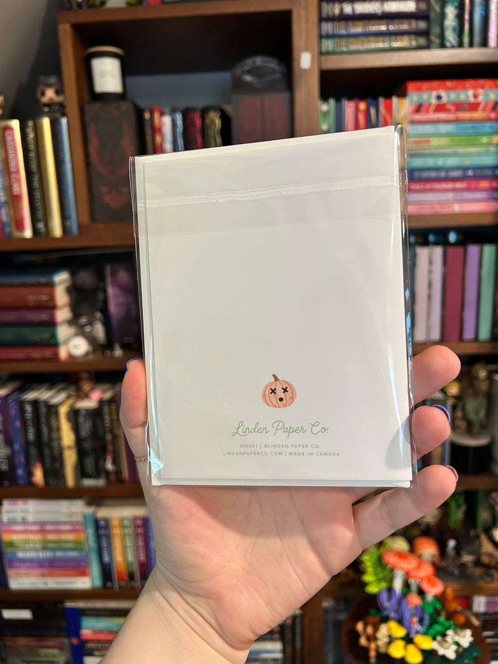 A hand holding up the back of a greeting card, featuring a small pink pumpkin illustration and the text 'Linden Paper Co' on the bottom, with a blurred bookshelf background.