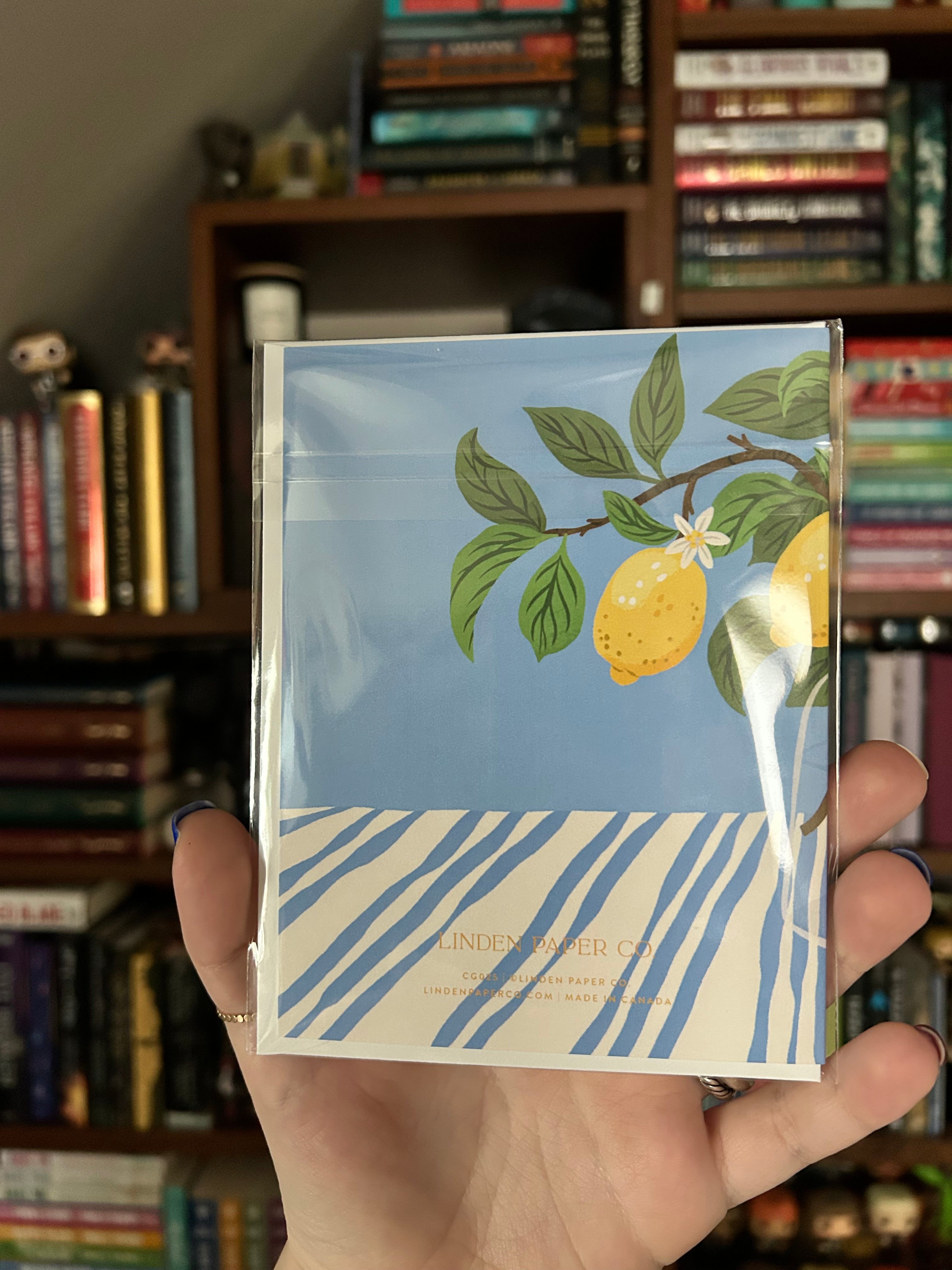A hand holding a back of greeting card featuring a blue background with a lemon illustration and blue-and-white striped accents, set against a blurred background of bookshelves