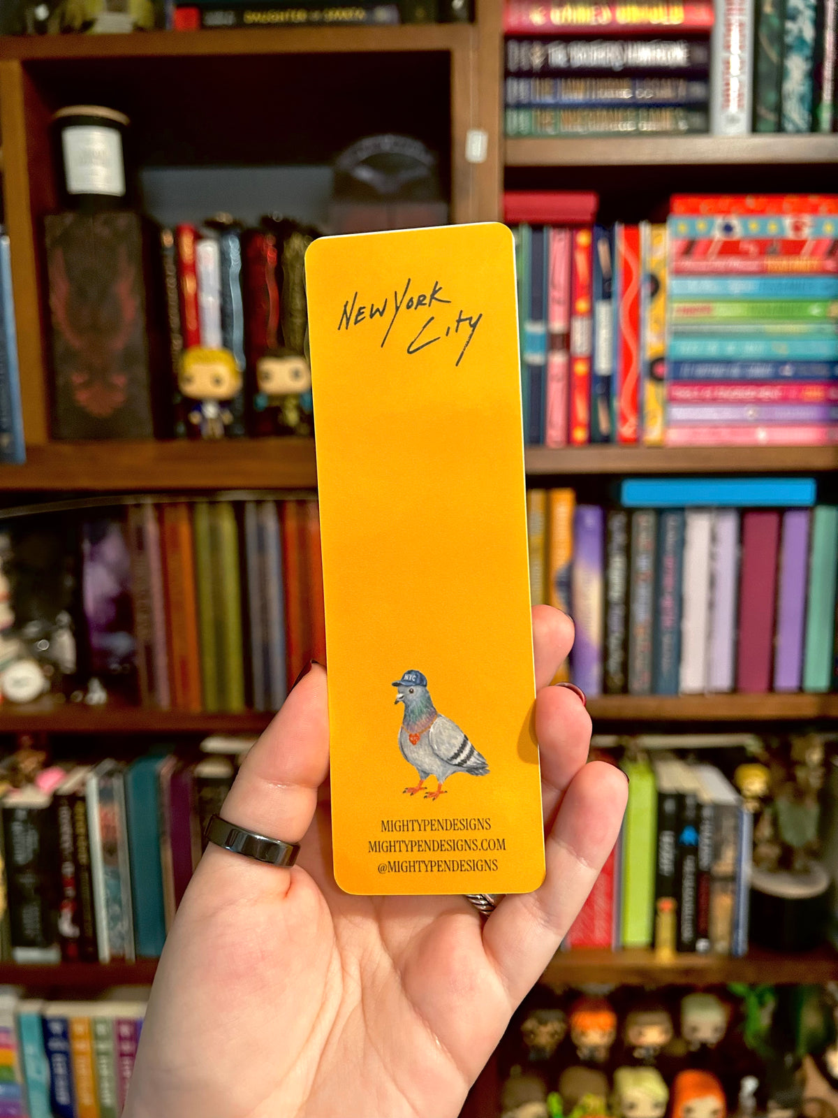 A hand holding a bright yellow bookmark featuring the words 'New York City' in black script and an illustration of a pigeon, with a blurred bookshelf background.