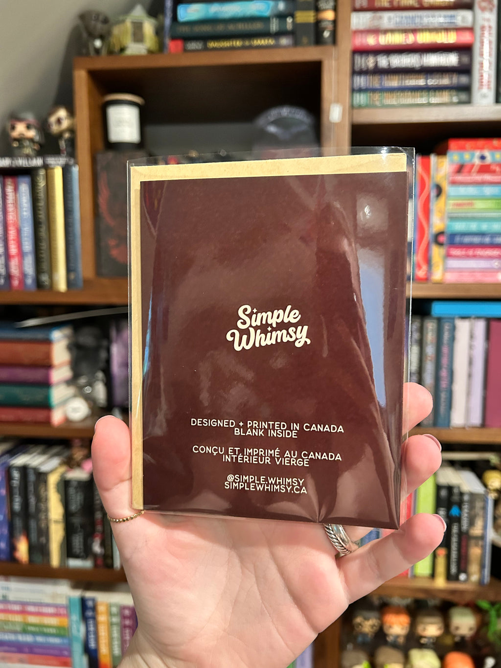 A hand holds a brown Simple Whimsy card or insert in a clear plastic sleeve, with a blurred bookshelf background.