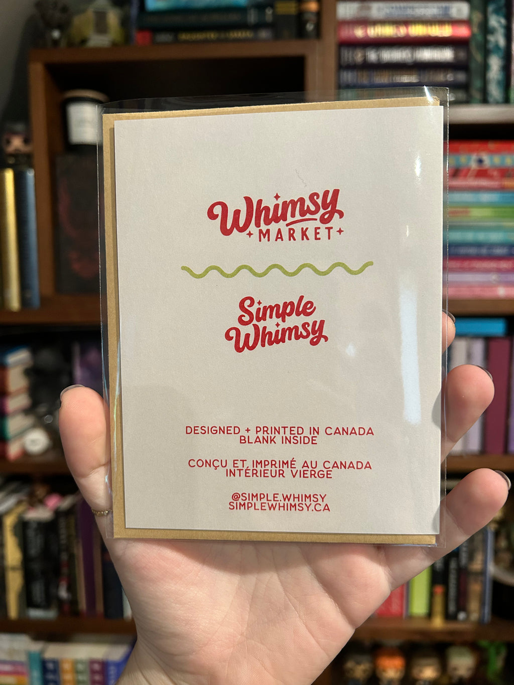 A hand holding a clear plastic sleeve displaying the back of a greeting card  with the text 'Whimsy Market' and 'Simple Whimsy' in red, photographed in front of a bookshelf.