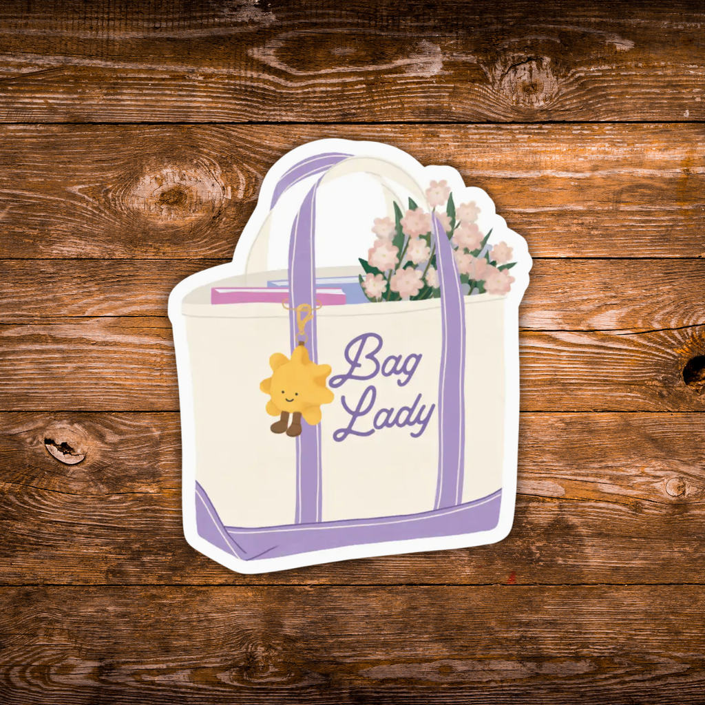 A die-cut sticker of a cream tote bag with lavender handles and trim, decorated with a yellow smiling sun charm, pink flowers, and the text 'Bag Lady'. The sticker is shown flat against a rustic brown wooden plank background.