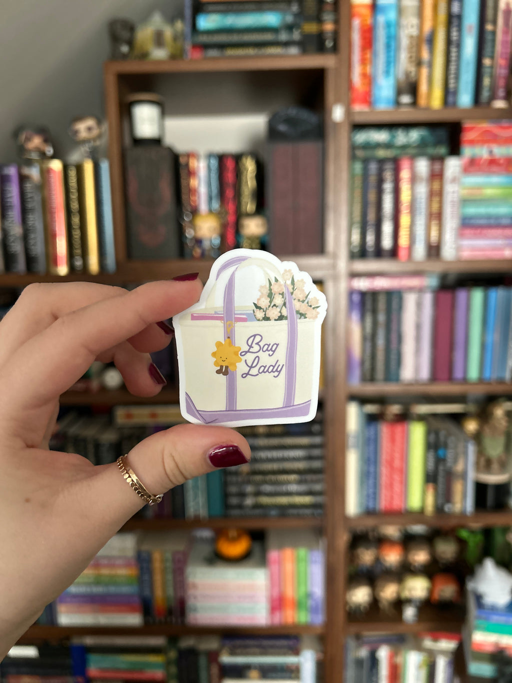 A hand with dark red nail polish holds a small decorative tag or sticker in front of a blurred bookshelf background. The tag is white with purple ribbon graphics, a small yellow sun, and the words 'Bag Lady' in purple script.