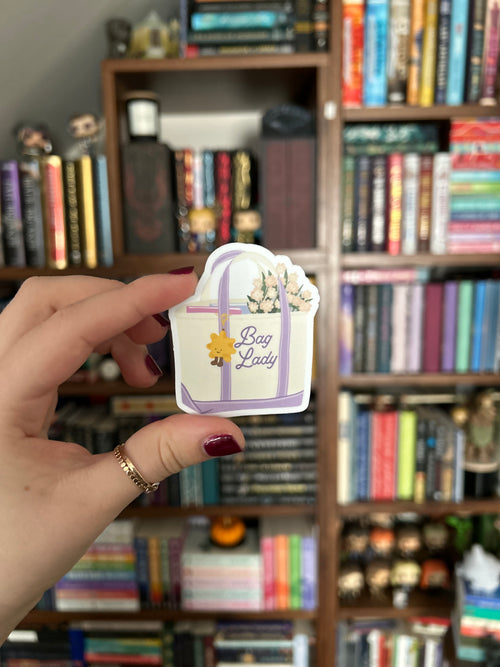 A hand with dark red nail polish holds a small decorative tag or sticker in front of a blurred bookshelf background. The tag is white with purple ribbon graphics, a small yellow sun, and the words 'Bag Lady' in purple script.