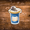 A die-cut sticker featuring a cartoon gray mouse lounging on a cup of coffee with an everything bagel on top, placed on a rustic brown wooden background. The sticker includes the text 'WE ARE HAPPY TO SERVE YOU'.
