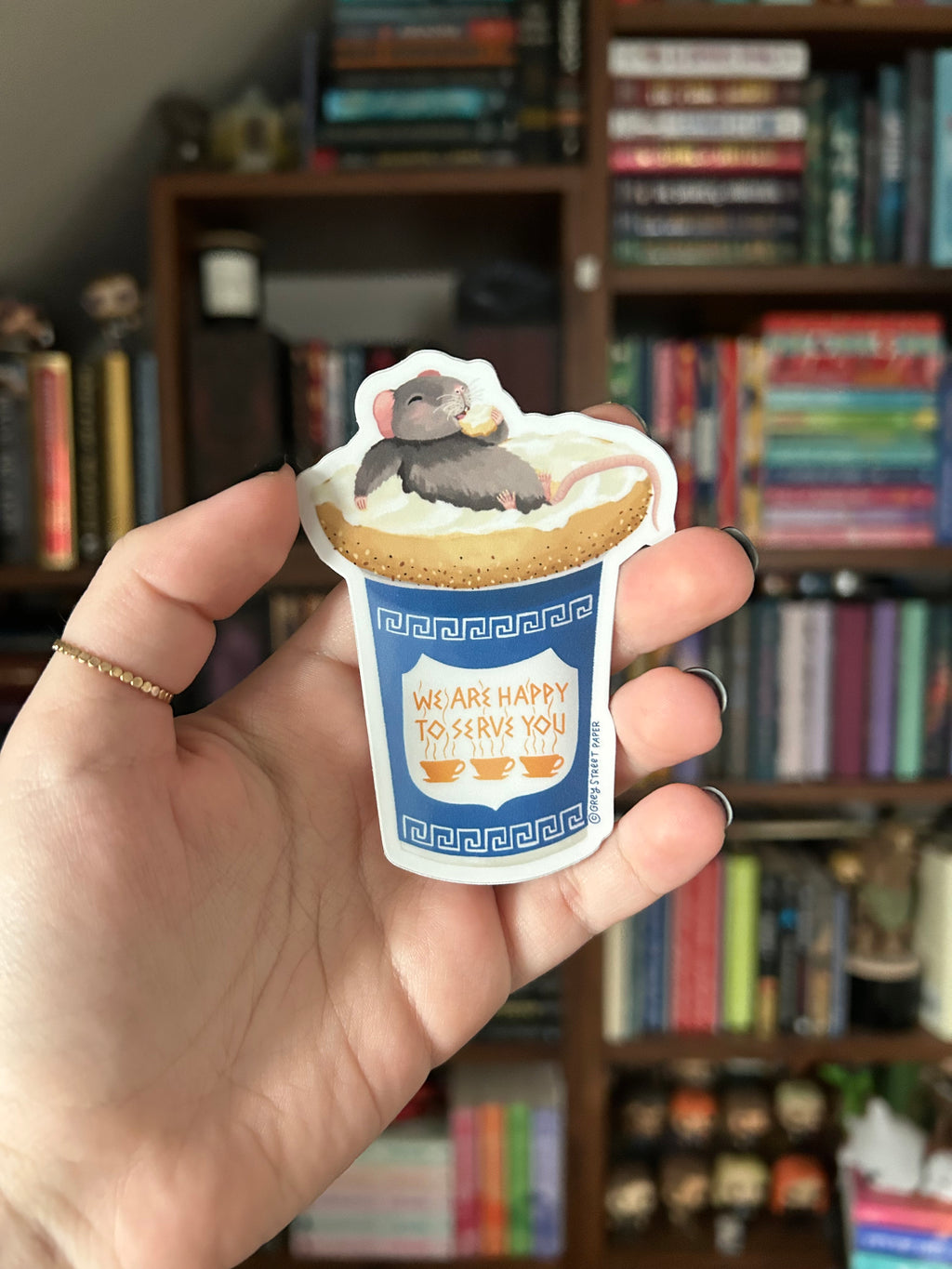 A hand holding a die-cut sticker featuring a cartoon mouse laying on everything bagel with cream cheese, with a blue label that reads 'WE ARE HAPPY TO SERVE YOU'. The background shows a blurred bookshelf.