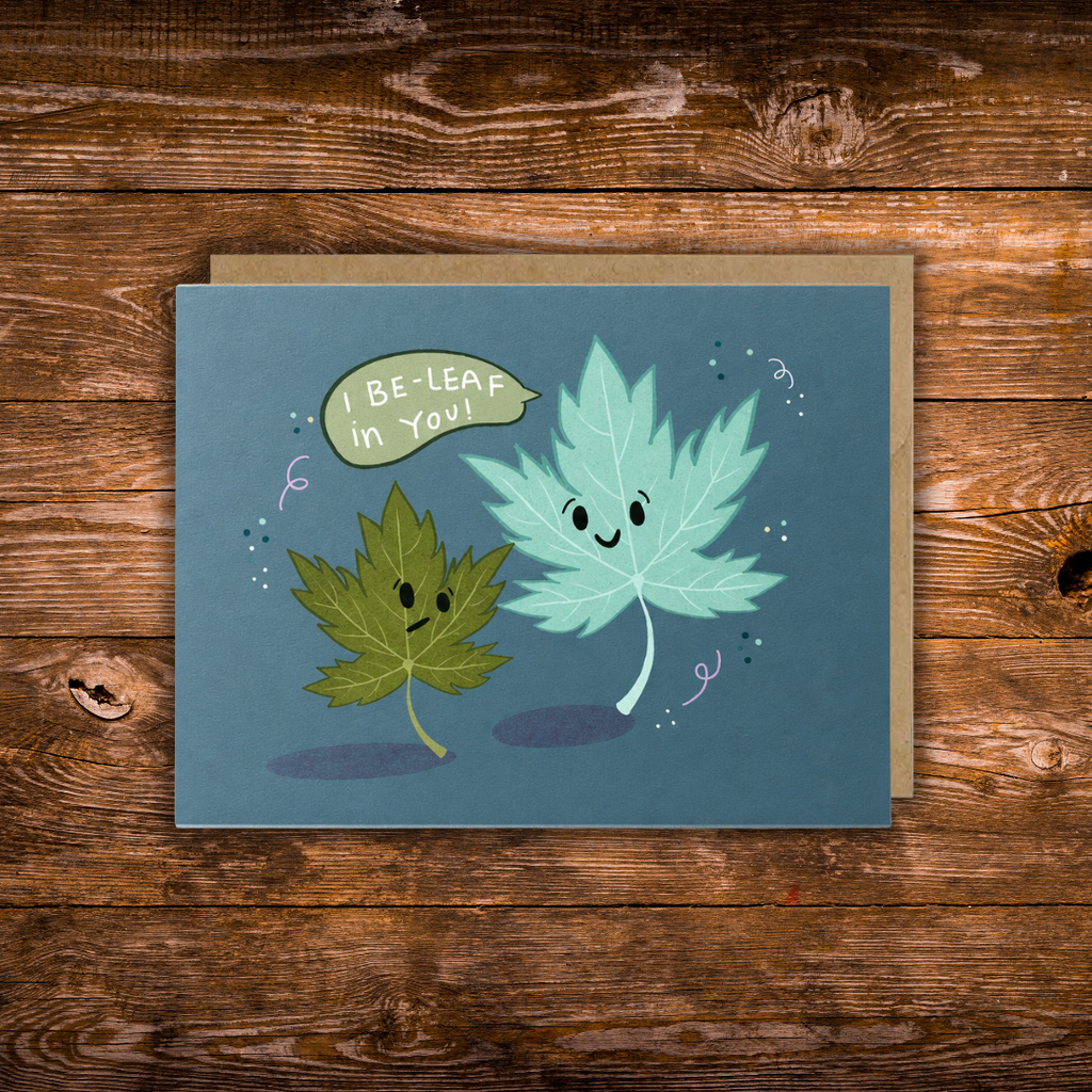 Landscape encouragement greeting card featuring two smiling illustrated leaves with the message "I Be-Leaf in You!" on a blue background.