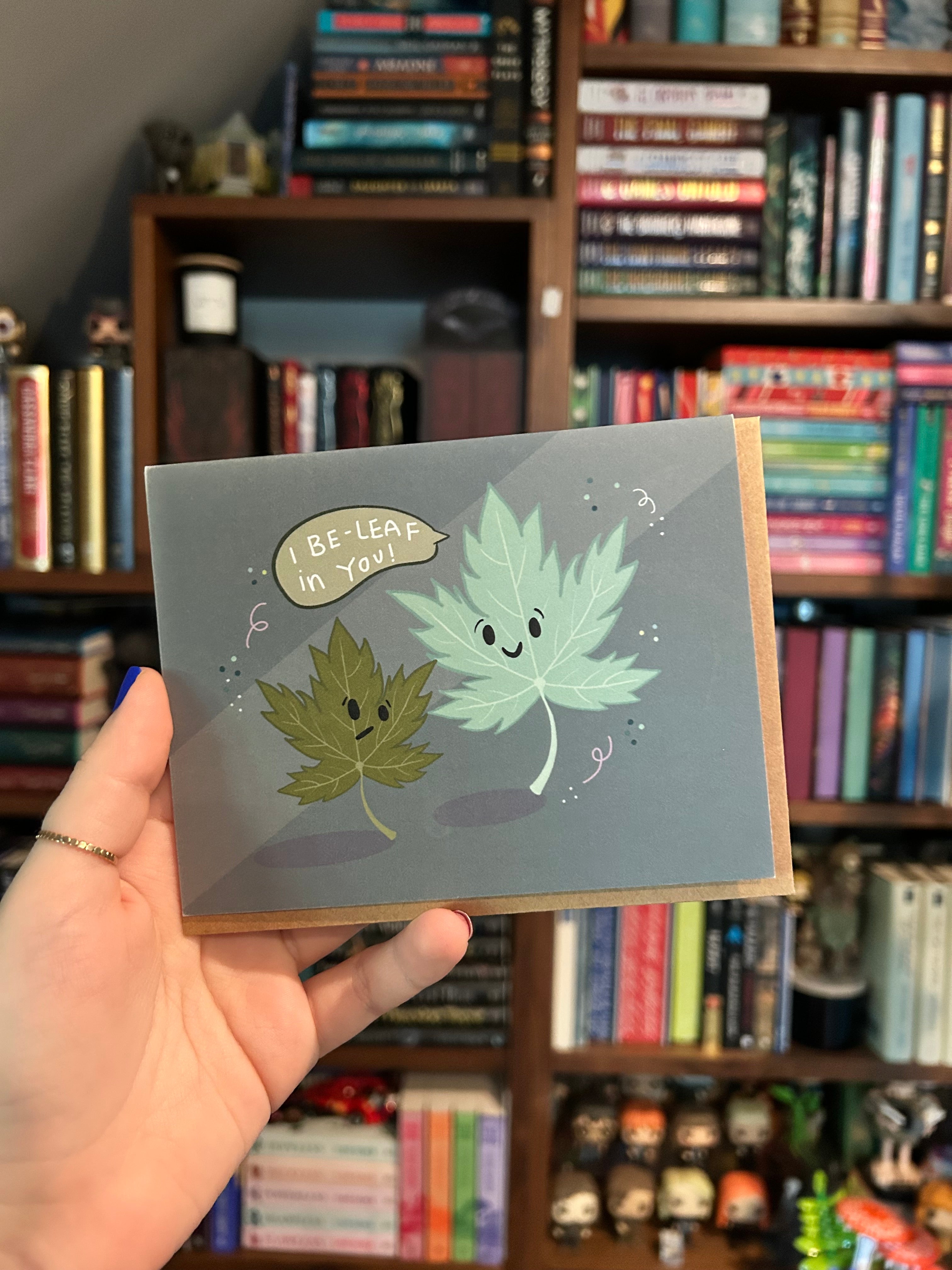 Landscape encouragement greeting card featuring two smiling illustrated leaves with the message "I Be-Leaf in You!" on a blue background.