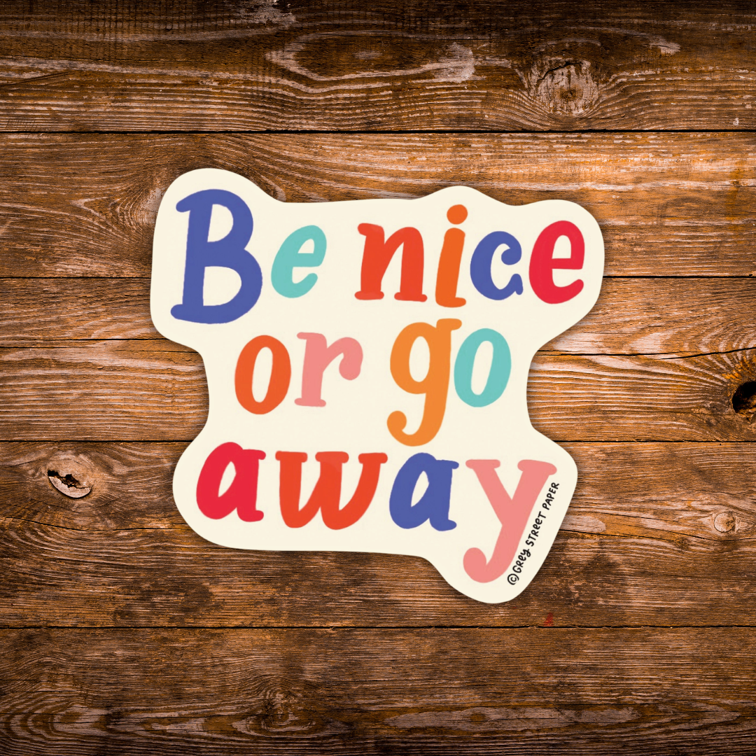 A die-cut sticker with the phrase 'Be nice or go away' in colorful retro-style lettering, placed on a rustic brown wooden surface. The sticker has a cream background and small text reading '©Grey Street Paper' along the bottom edge.