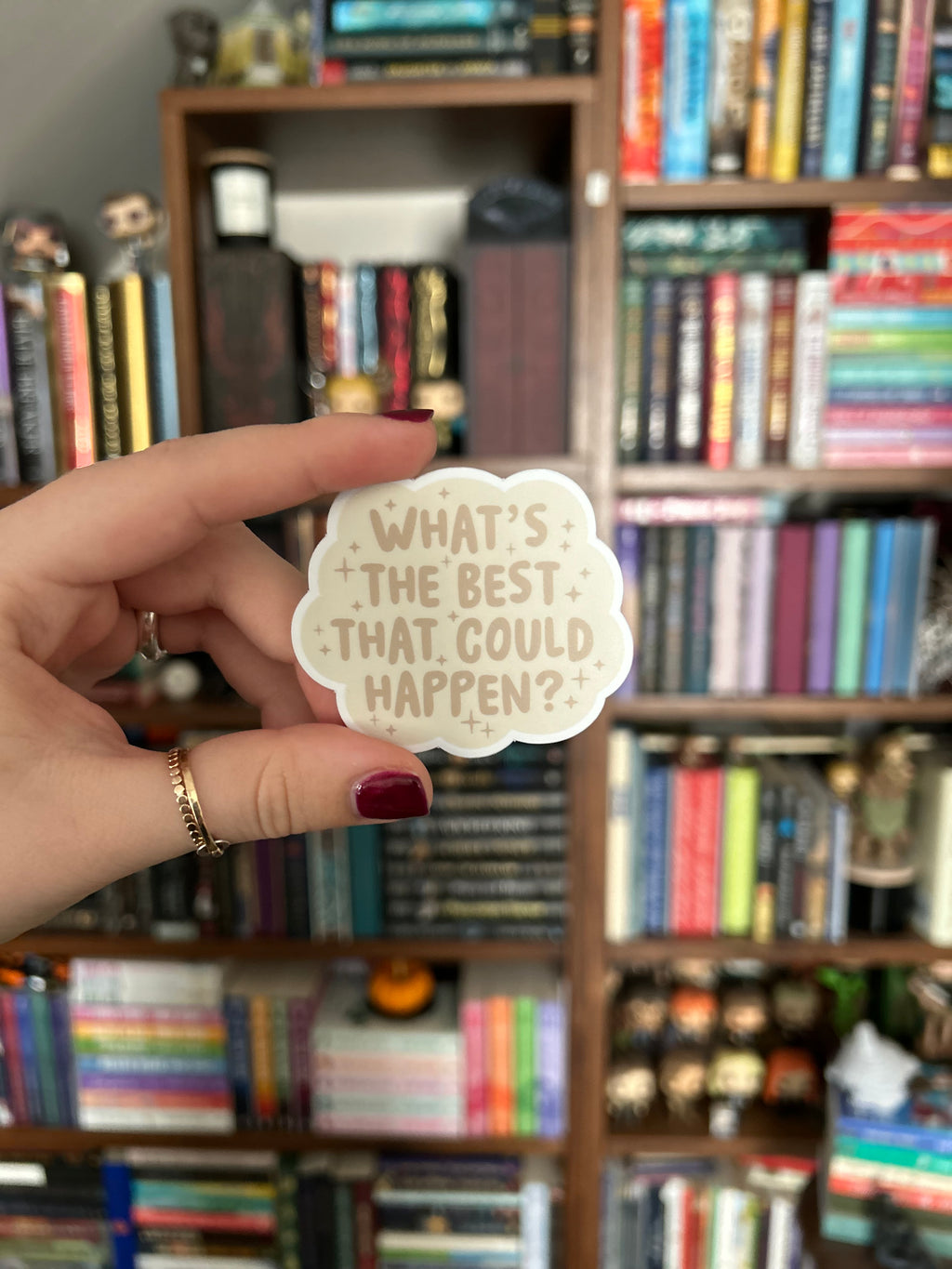 A hand with dark red nail polish holds a small, scalloped cream sticker with the text 'WHAT'S THE BEST THAT COULD HAPPEN?' in brown lettering, set against a blurred background of bookshelves filled with books.