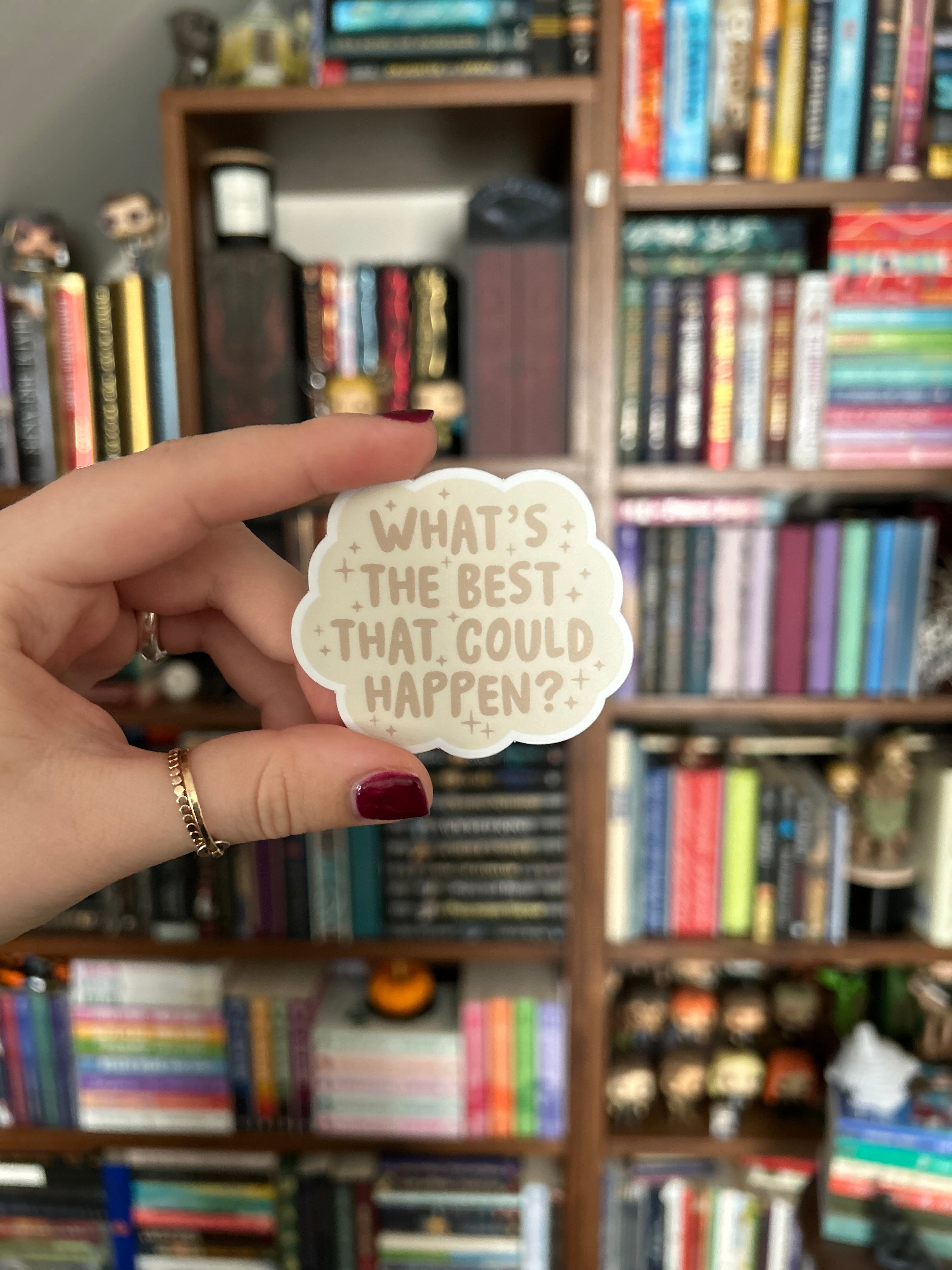 A hand with dark red nail polish holds a small, scalloped cream sticker with the text 'WHAT'S THE BEST THAT COULD HAPPEN?' in brown lettering, set against a blurred background of bookshelves filled with books.