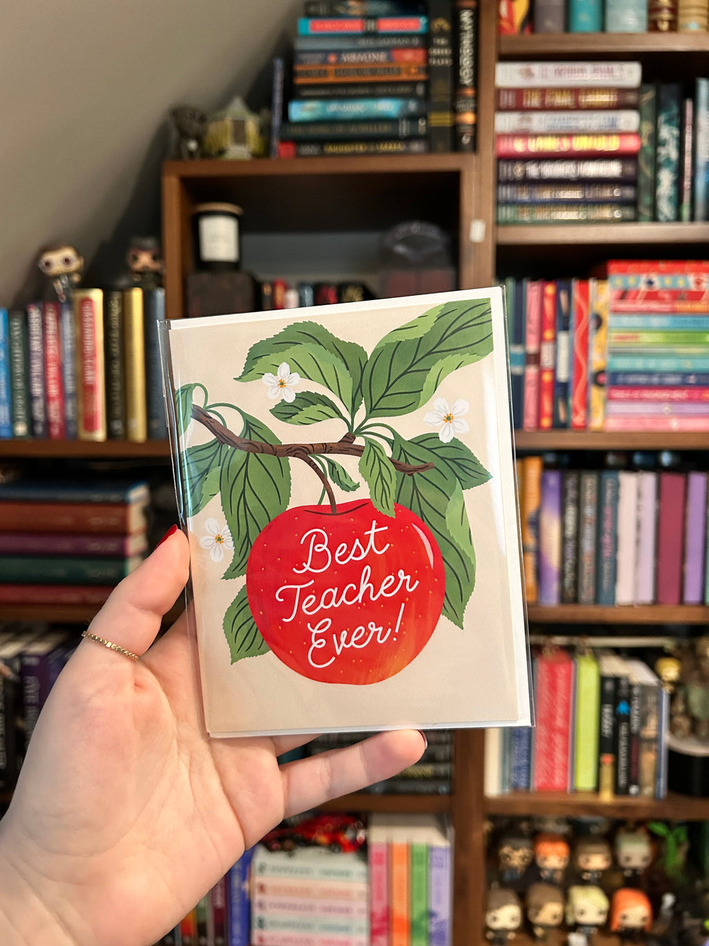 Teacher appreciation greeting card featuring a bright red apple hanging from a leafy branch with white blossoms and the words "Best Teacher Ever!" in white script.