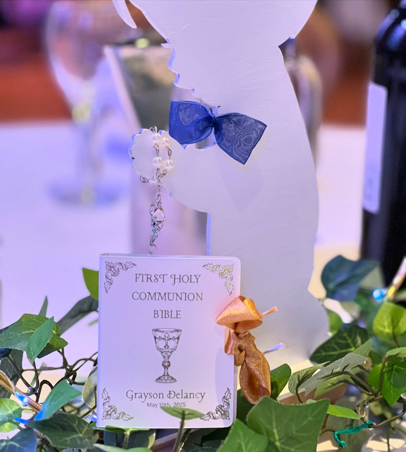 A personalized first communion Tic Tac Bible, decorated with a small silver chalice and a gold ribbon, displayed on a table with green ivy and a white communion silhouette backdrop.