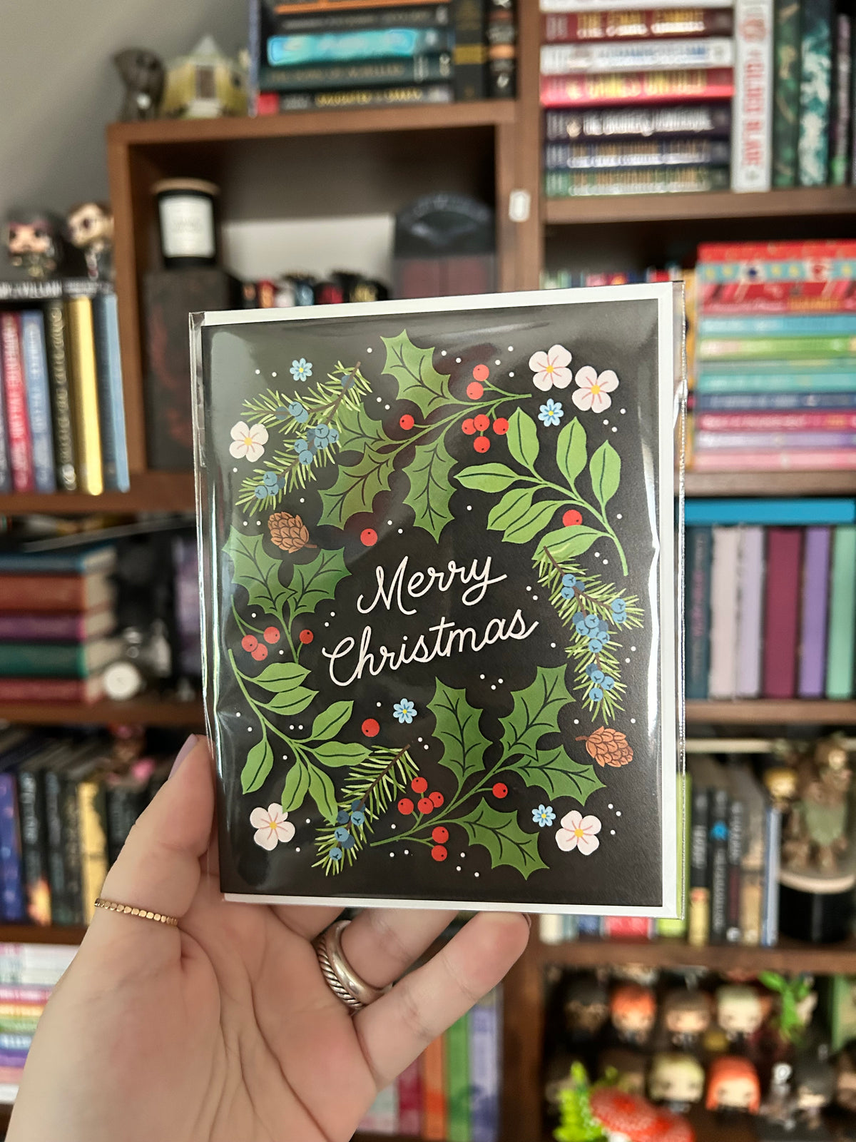 A hand holds a Christmas greeting card with a black background and a colorful holly and berry wreath design, with the words 'Merry Christmas' in white script. The background shows a blurred bookshelf filled with books.