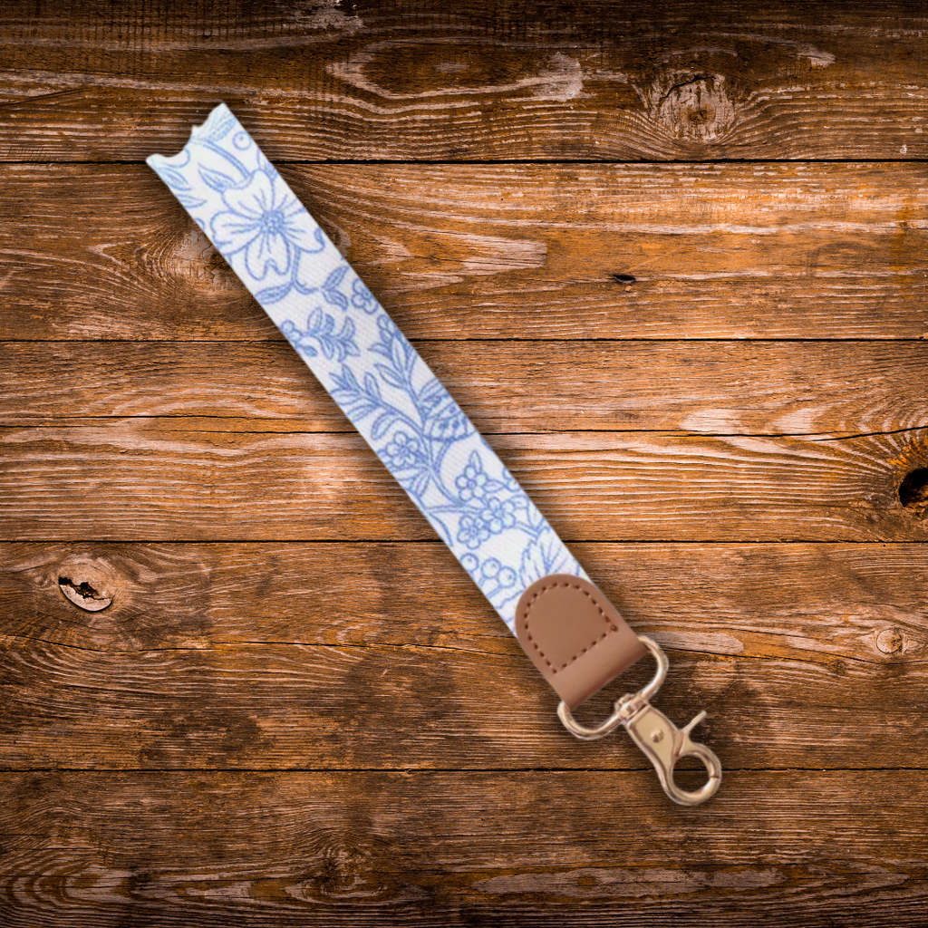 A fabric wrist strap with a light blue and white floral pattern, finished with a brown leather tab and a gold-tone metal clasp, displayed diagonally on a rustic wooden plank background.