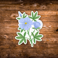 A die-cut sticker featuring light blue flowers and green leaves, displayed on a rustic brown wooden surface.
