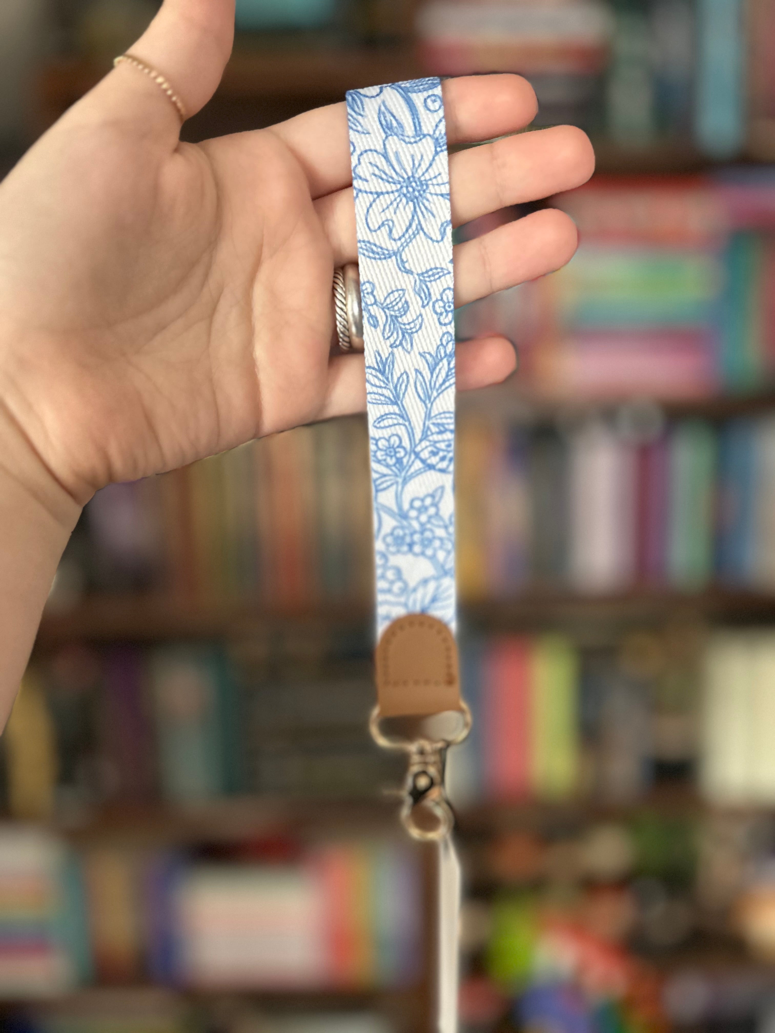 A close-up of a hand holding a fabric wrist strap with a blue floral pattern on a white background, featuring a tan leather loop and a gold metal clasp, with a blurred bookshelf in the background.