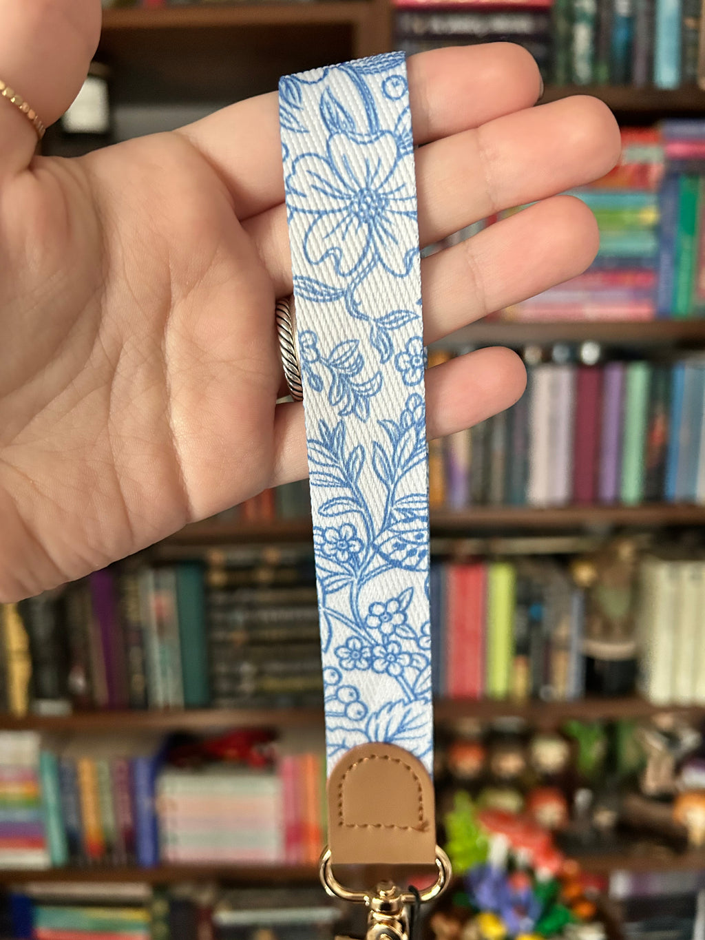 A close-up of a hand holding a fabric wrist strap with a blue floral pattern on a white background, featuring a tan leather tab and gold-tone hardware, with a blurred bookshelf in the background.