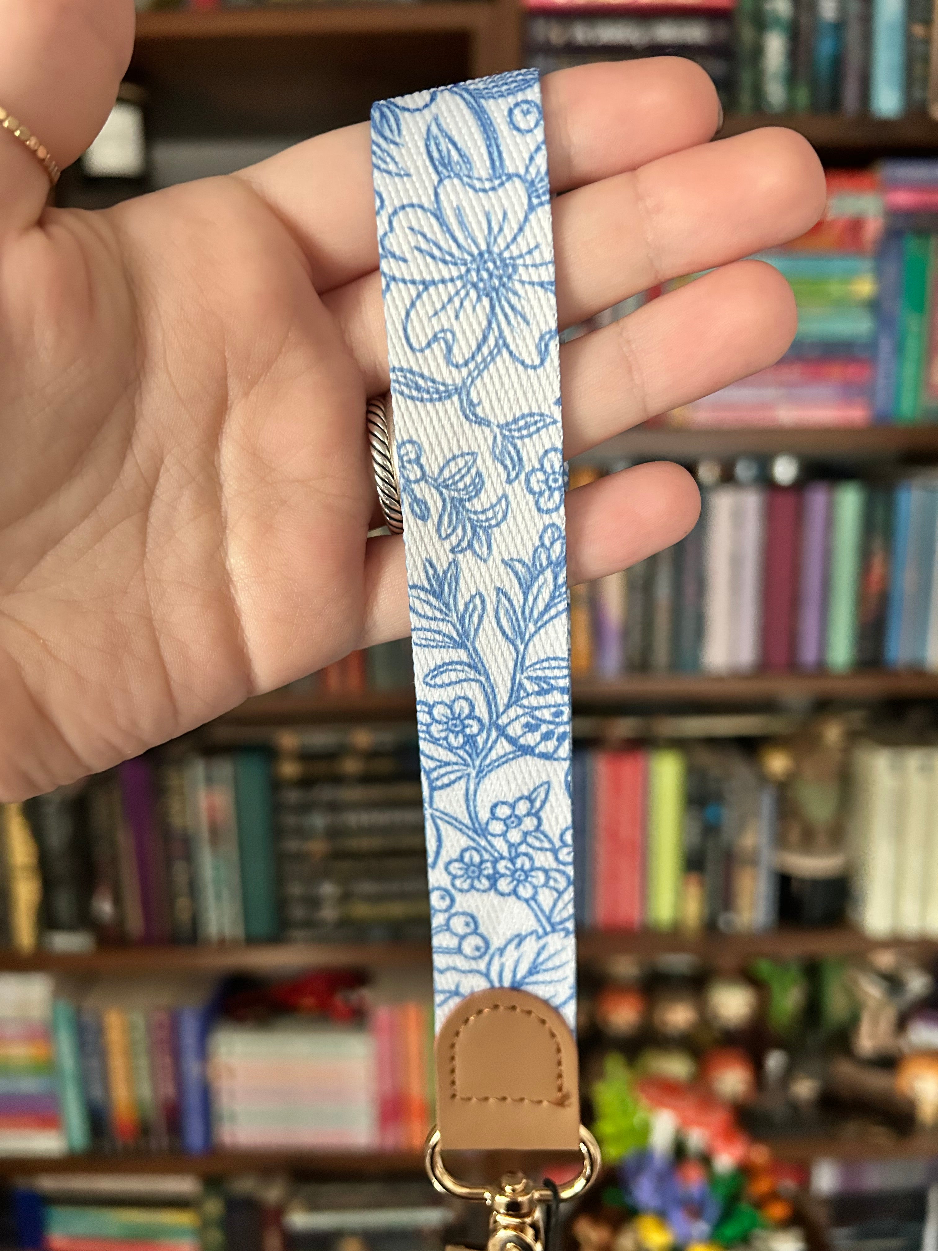 A close-up of a hand holding a fabric wrist strap with a blue floral pattern on a white background, featuring a tan leather tab and gold-tone hardware, with a blurred bookshelf in the background.