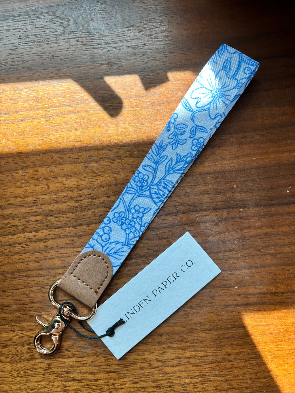 A fabric wrist strap with a blue floral pattern on a white background, finished with a tan leather tab and gold-tone clasp, shown on a wooden table with a paper tag reading 'LINDEN PAPER CO.'.