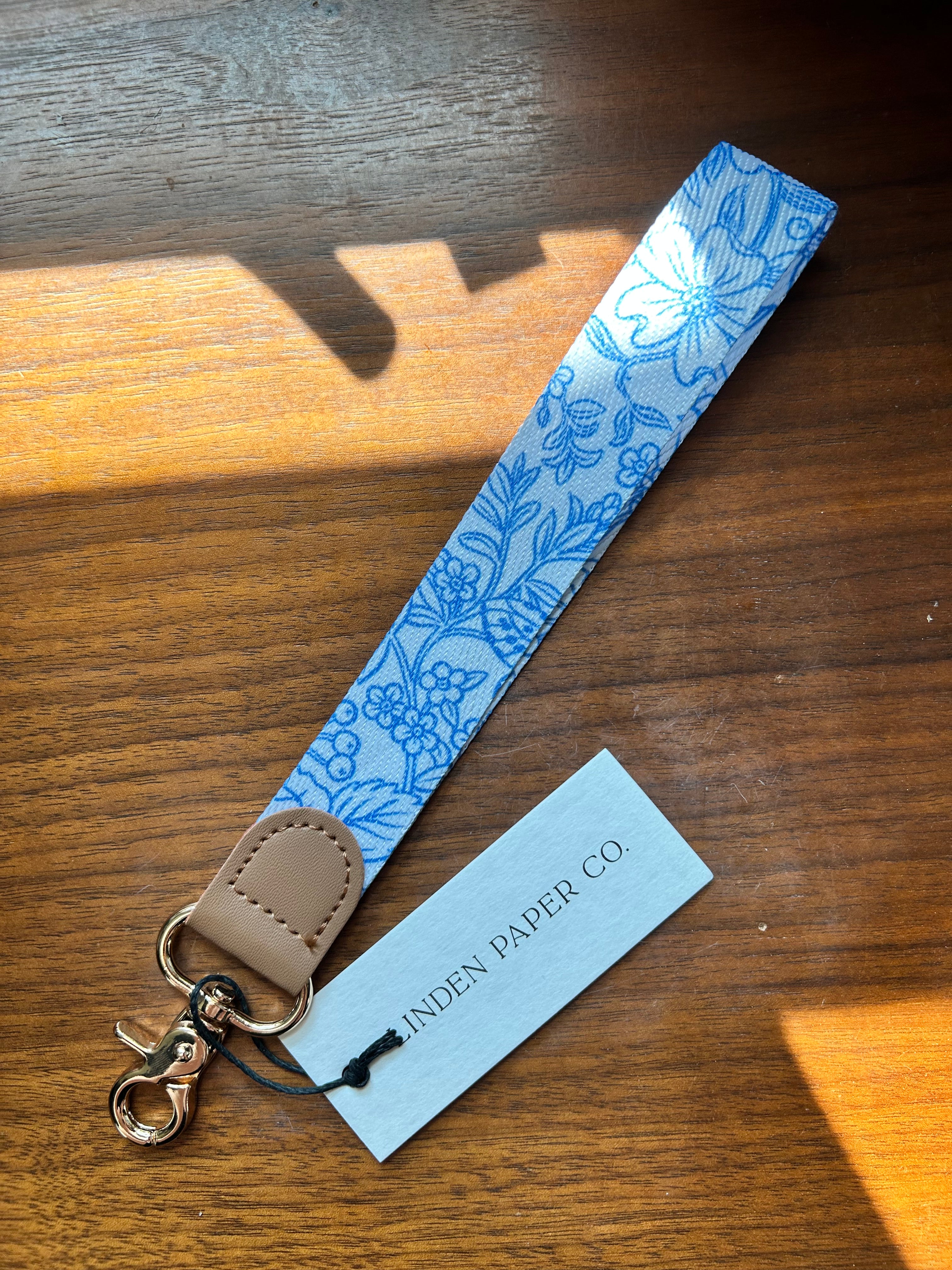 A fabric wrist strap with a blue floral pattern on a white background, finished with a tan leather tab and gold-tone clasp, shown on a wooden table with a paper tag reading 'LINDEN PAPER CO.'.