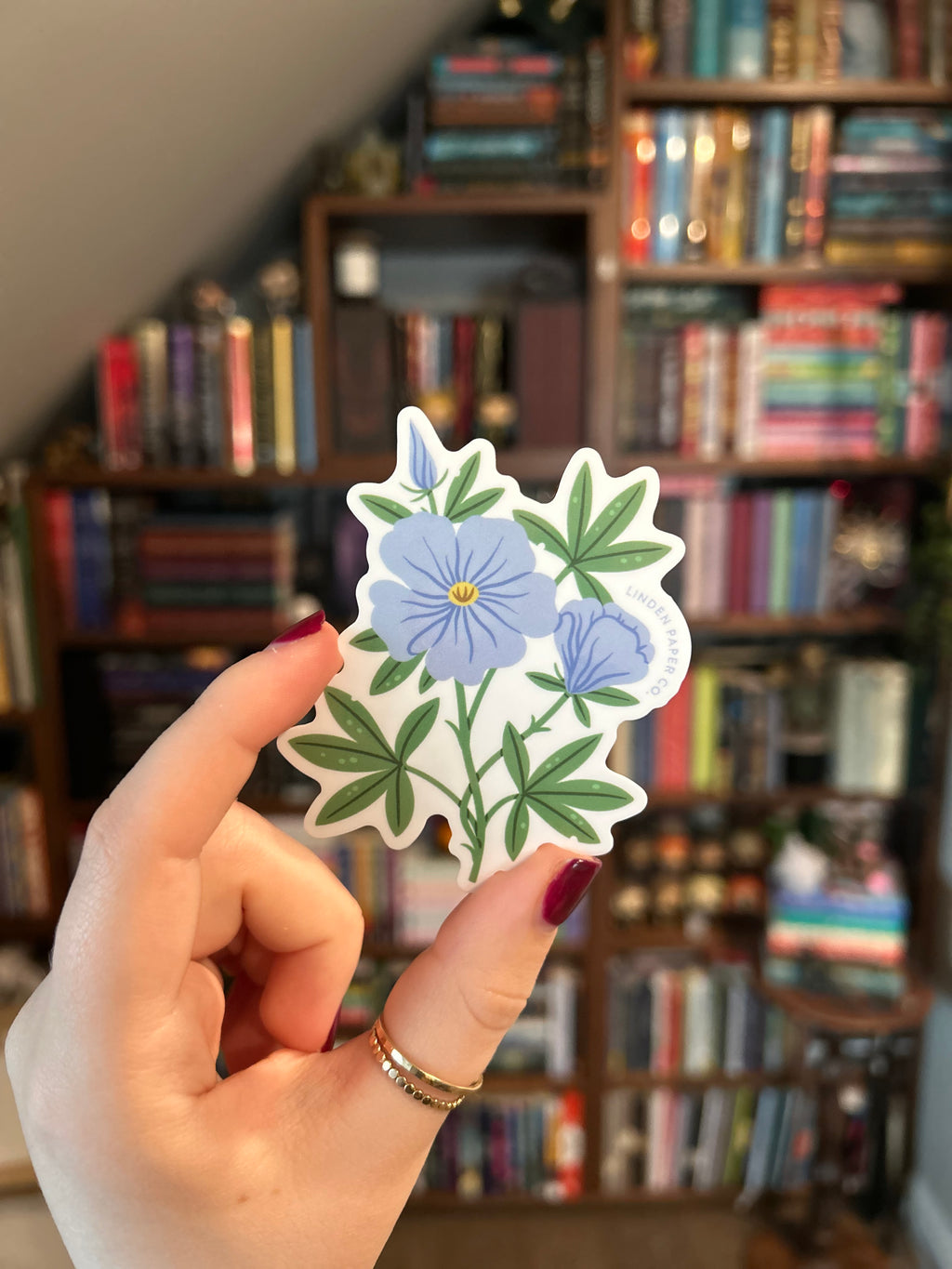 A hand with dark red nail polish holds a die-cut sticker featuring blue flowers and green leaves in front of a blurred bookshelf background.
