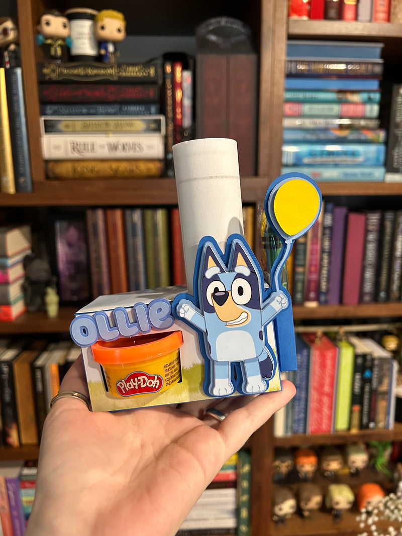 A hand holds a custom Bluey-themed Play-Dah set in front of a bookshelf. The set includes a coloring pages, a small orange tub of Play-Dah, a cardboard box with 'Bluey' and 'Play-Doh' branding, and a blue balloon