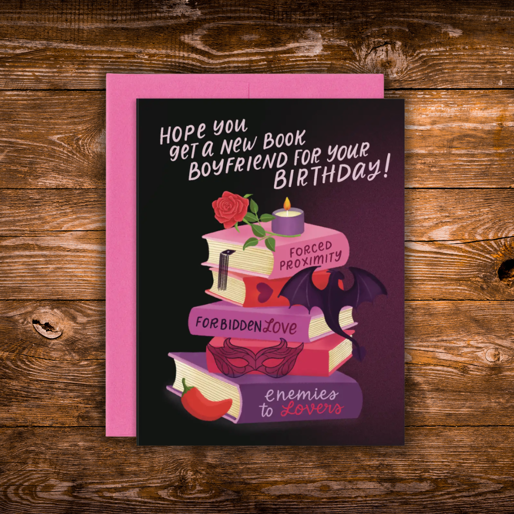 A birthday card with a pink envelope, featuring a dark purple and black background with an illustration of stacked books, a candle, a rose, a bat, and a red pepper. The text reads, 'HOPE YOU GET A NEW BOOK BOYFRIEND FOR YOUR BIRTHDAY!'.