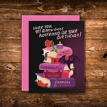 A birthday card with a pink envelope, featuring a dark purple and black background with an illustration of stacked books, a candle, a rose, a bat, and a red pepper. The text reads, 'HOPE YOU GET A NEW BOOK BOYFRIEND FOR YOUR BIRTHDAY!'.