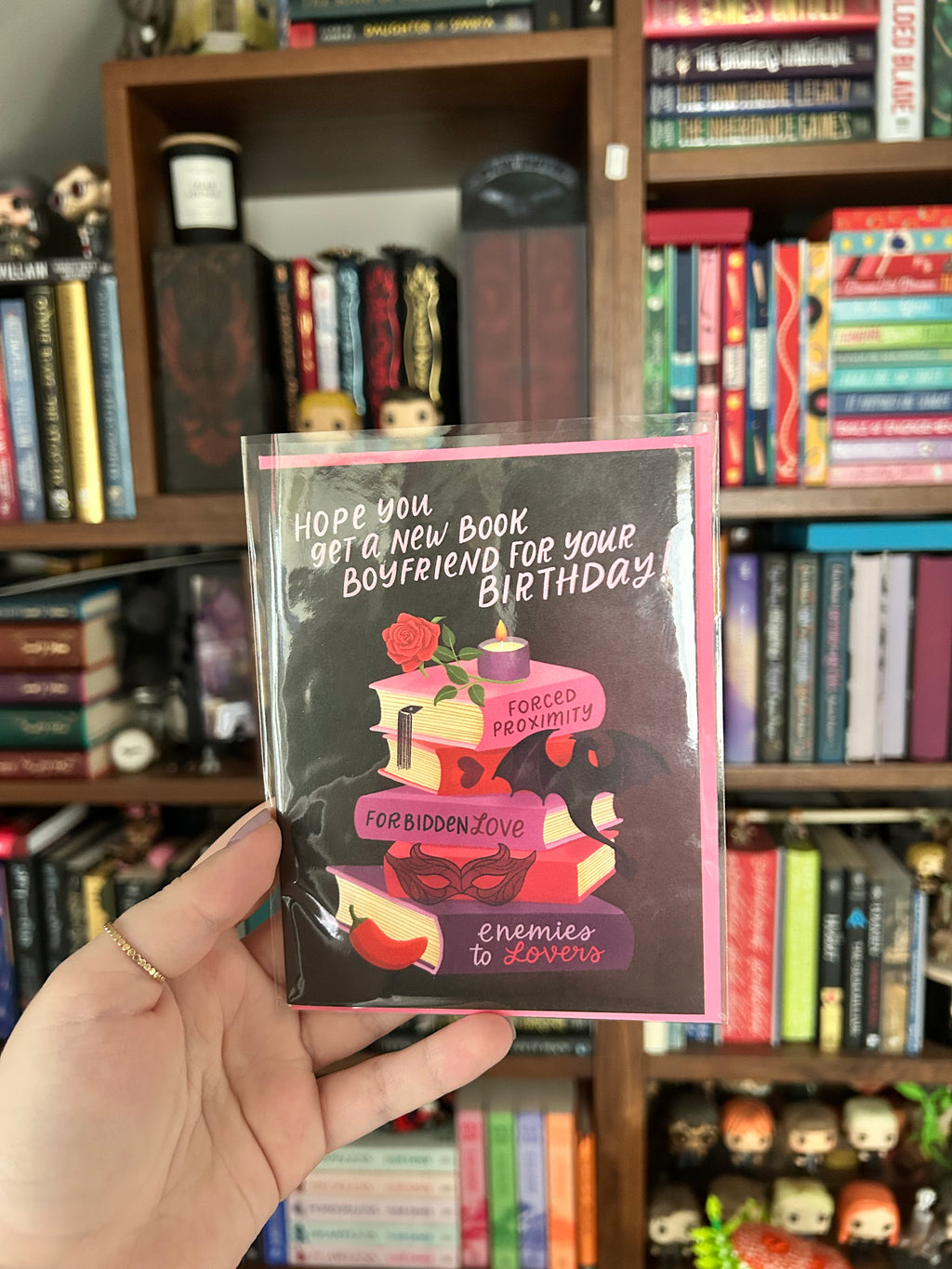 A hand holds a square greeting card with a pink border and a dark illustration featuring stacked books, a rose, and bold text that reads, 'Hope you get a new book for your boyfriend your birthday! Forced Proximity Forbidden Love Enemies to Enemies.' The background shows a blurred bookshelf filled with books.