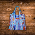 A reusable tote bag with a light blue base and a colorful collage print featuring glasses, books, and text, laid flat on a rustic wooden plank background.
