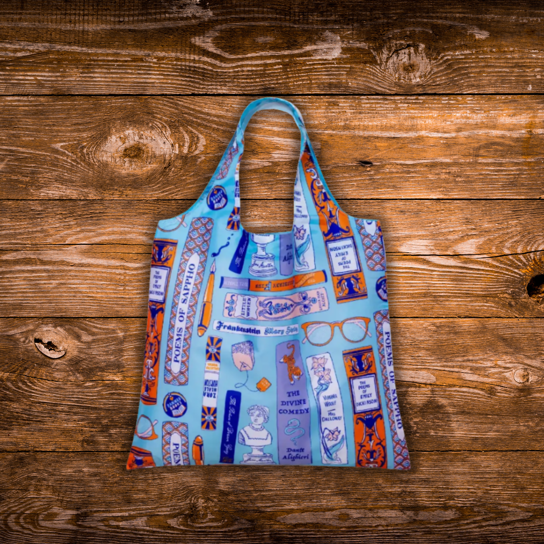 A reusable tote bag with a light blue base and a colorful collage print featuring glasses, books, and text, laid flat on a rustic wooden plank background.