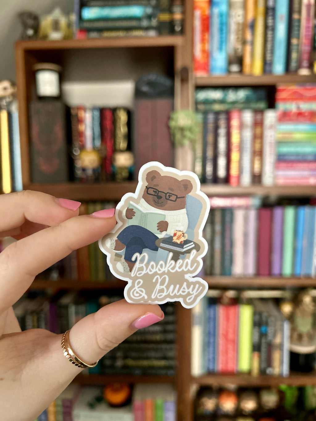 A hand with pink nail polish holds a die-cut sticker featuring a cartoon bear wearing glasses and reading, with the text 'Booked & Busy' in white script. The background is a blurred bookshelf filled with books.