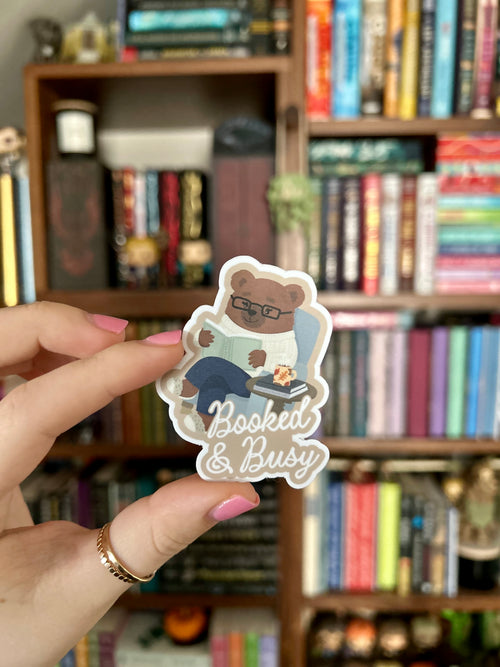 A hand with pink nail polish holds a die-cut sticker featuring a cartoon bear wearing glasses and reading, with the text 'Booked & Busy' in white script. The background is a blurred bookshelf filled with books.