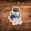 A die-cut sticker featuring an illustrated brown bear wearing glasses and a white sweater, sitting in a chair and reading a book with the words 'Booked & Busy' in white script. The sticker is displayed on a rustic brown wooden background.