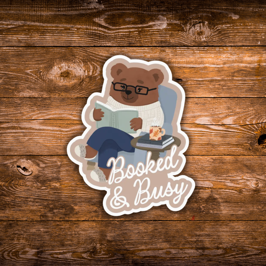 A die-cut sticker featuring an illustrated brown bear wearing glasses and a white sweater, sitting in a chair and reading a book with the words 'Booked & Busy' in white script. The sticker is displayed on a rustic brown wooden background.